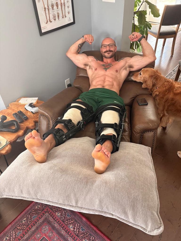 Brian flexing in a recliner with braces on both knees after surgery, his dog beside him